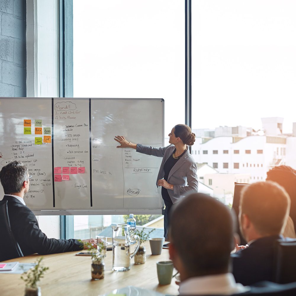 A professional female strategist from Planify Agency leads a collaborative marketing strategy session in a sun-drenched, modern boardroom. Standing before a large whiteboard filled with project notes, content plans, and sticky notes, she gestures toward a "Reporting" section while presenting to a team of professionals. The large windows in the background offer an expansive city view, emphasizing the high-level, forward-thinking approach of Planify Agency’s Marketing Strategy services.